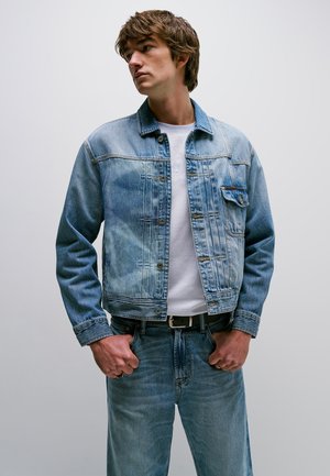 Jonge man die een verweerde blauwe spijkerjas draagt over een wit T-shirt en bijpassende blauwe jeans, staand met handen dicht bij de riem tegen een effen achtergrond.