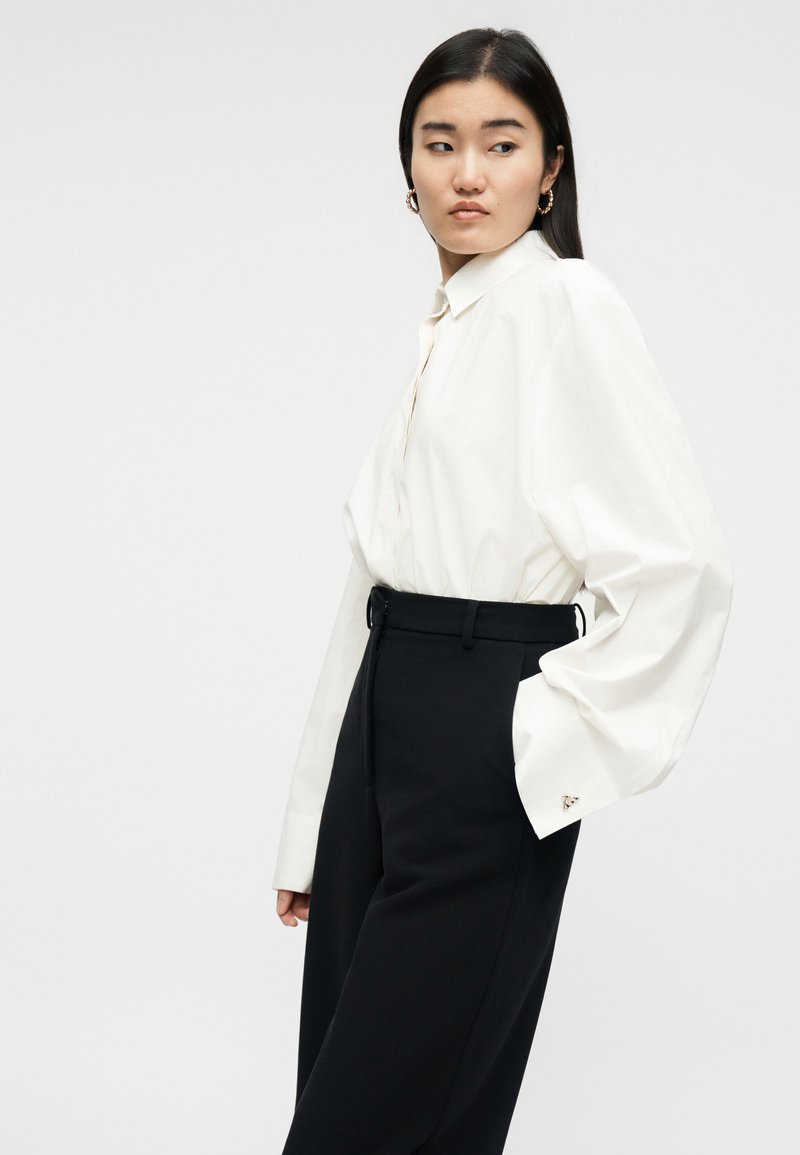 Frau mit langen schwarzen Haaren, die goldene Creolen, ein weißes, übergroßes Hemd und hoch taillierte schwarze Hosen trägt, schaut seitlich.