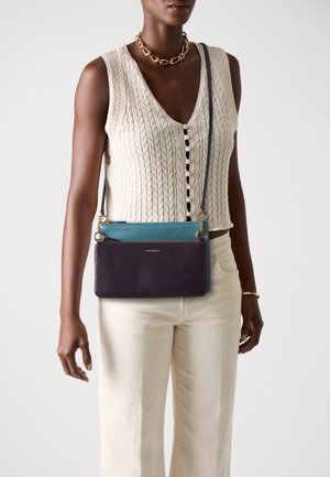 Woman wearing cream cable-knit sleeveless top, cream pants, gold necklace, and carrying two leather crossbody bags in teal and dark purple.
