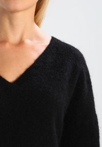 Schwarzer, gestrickter Pullover mit V-Ausschnitt, der eine weiche, flauschige Textur und eine lockere Passform aufweist. Der Stoff wirkt warm und gemütlich.