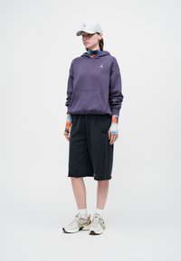 Sudadera con capucha morada y bolsillo delantero, pantalones cortos negros a la altura de la rodilla y gorra gris claro. Zapatillas blancas y beige con acentos en la entresuela.