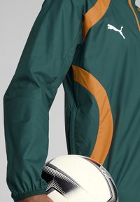 Green sports jacket with orange accents, featuring a zipped collar and elastic cuffs. A white soccer ball with black designs is held in front.