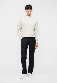 Άνδρας που στέκεται μπροστά σε λευκό φόντο, φορώντας ανοιχτό μπεζ φούτερ, μαύρο παντελόνι και λευκά αθλητικά παπούτσια, με το ένα χέρι στην τσέπη.