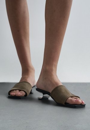 Voeten met bruine open-toe muilen met gebogen lage hakken op een grijze vloer tegen een lichte achtergrond.