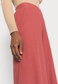 Pantalons larges à côtes rouges avec une taille haute, associés à un pull ajusté beige, mettant en valeur une texture lisse et des lignes épurées.