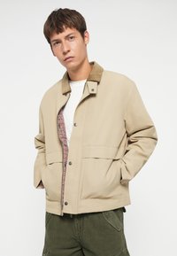 Beige jacket with a corduroy collar, snap buttons, and a checkered lining. Front pockets, smooth texture, and relaxed fit.