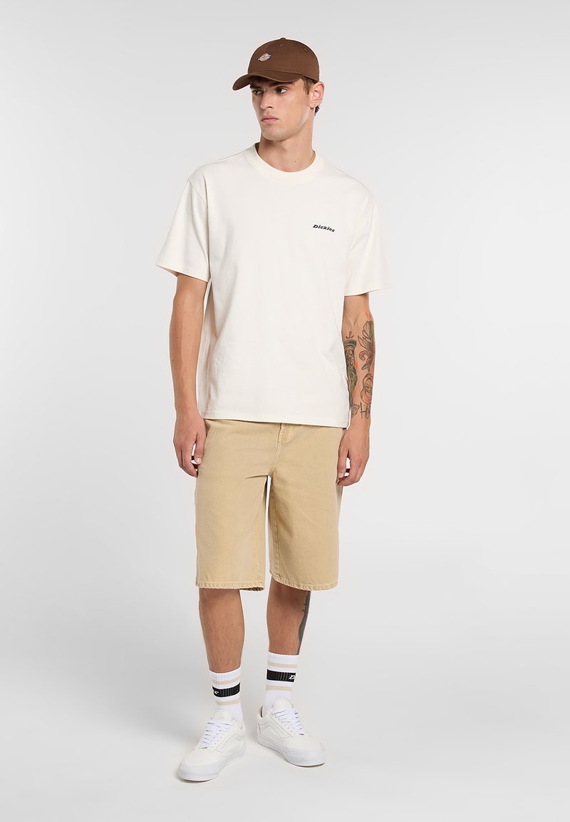 Camiseta de algodón beige con mangas cortas y logo negro, combinada con pantalones cortos de mezclilla beige. Zapatillas blancas y gorra marrón completan el conjunto.