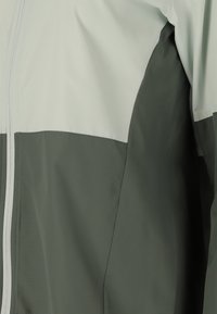 Veste imperméable avec un design bicolore, comportant une section supérieure gris clair et une section inférieure gris foncé. Elle est dot�ée d'une fermeture éclair intégrale et d'une texture lisse.