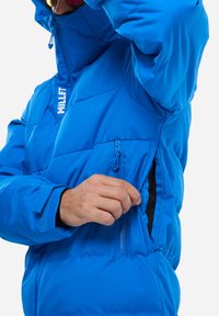 Blaue isolierte Jacke mit hohem Kragen, Reißverschlusstasche, verstellbaren Manschetten und einem gesteppten Design für Wärme. Minimale Logo-Detaillierung.