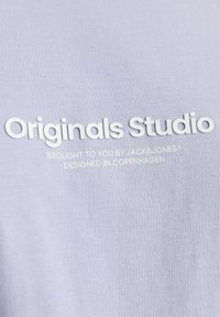 Šviesiai violetinė marškinė su lygiu paviršiumi, kuriose išryškinta balta spauda: "Originals Studio" ir mažesnis tekstas žemiau.