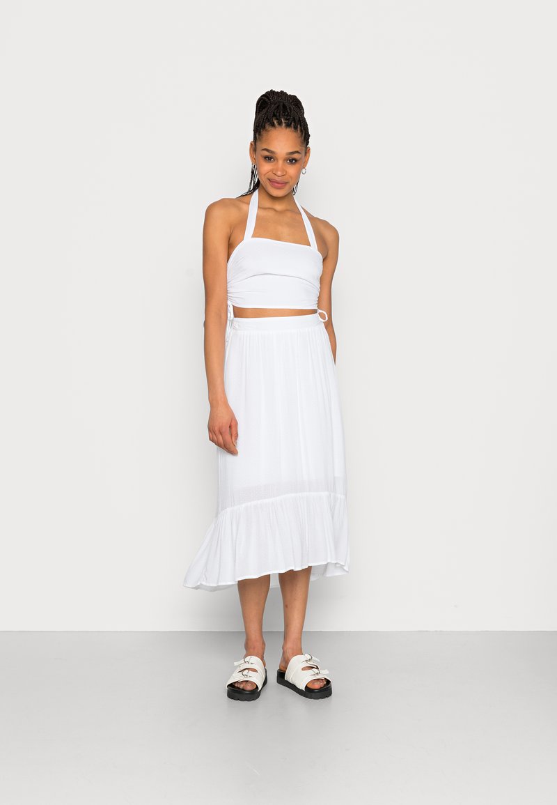 Conjunto branco de duas peças, apresentando um top curto com decote halter e uma saia midi fluida com barra frisada. Usado com sandálias brancas.