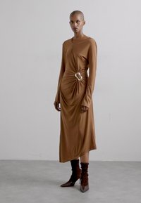 Vestido marrón de manga larga hecho de una tela suave, con un diseño retorcido en la cintura, acentuado con un detalle de anillo dorado.