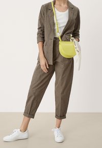 Completo blazer e pantaloni con motivo a occhio di pernice marrone, abbinato a una maglietta bianca a costine, sneaker bianche e una piccola borsa a tracolla gialla.