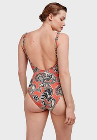 Eén delig badpak in koraal met zwart bloemenpatroon, heeft brede banden en een lage rugontwerp, gemaakt van rekbare stof voor comfort.
