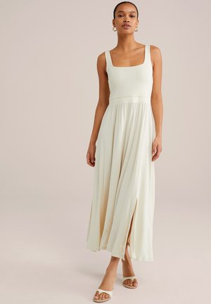 Cream maxi dress with a square neckline, wide straps, and pleated skirt featuring side slits. Worn with minimal sandals.