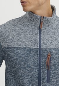 Textūruotas pūkų džemperis su tamsiai mėlynos ir šviesiai pilkos spalvų blokuoto dizaino, aukštu apykakle ir užtrauktuku užsegama kišene su rudos odos akcentu.