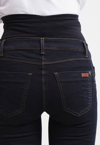 Jeans de cintura alta azul-escuro com um cós elástico, costuras visíveis e dois bolsos traseiros com uma pequena etiqueta de marca castanha.