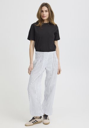 Femme debout portant un t-shirt noir à manches courtes, un pantalon blanc taille haute à rayures verticales noires, et des baskets beiges avec des rayures noires.