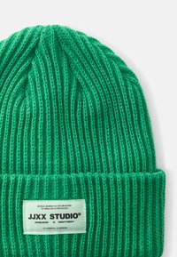 Beanie verde a maglia con texture a coste e risvolto. Presenta un'etichetta rettangolare con il nome del marchio e testo in nero su uno sfondo chiaro.