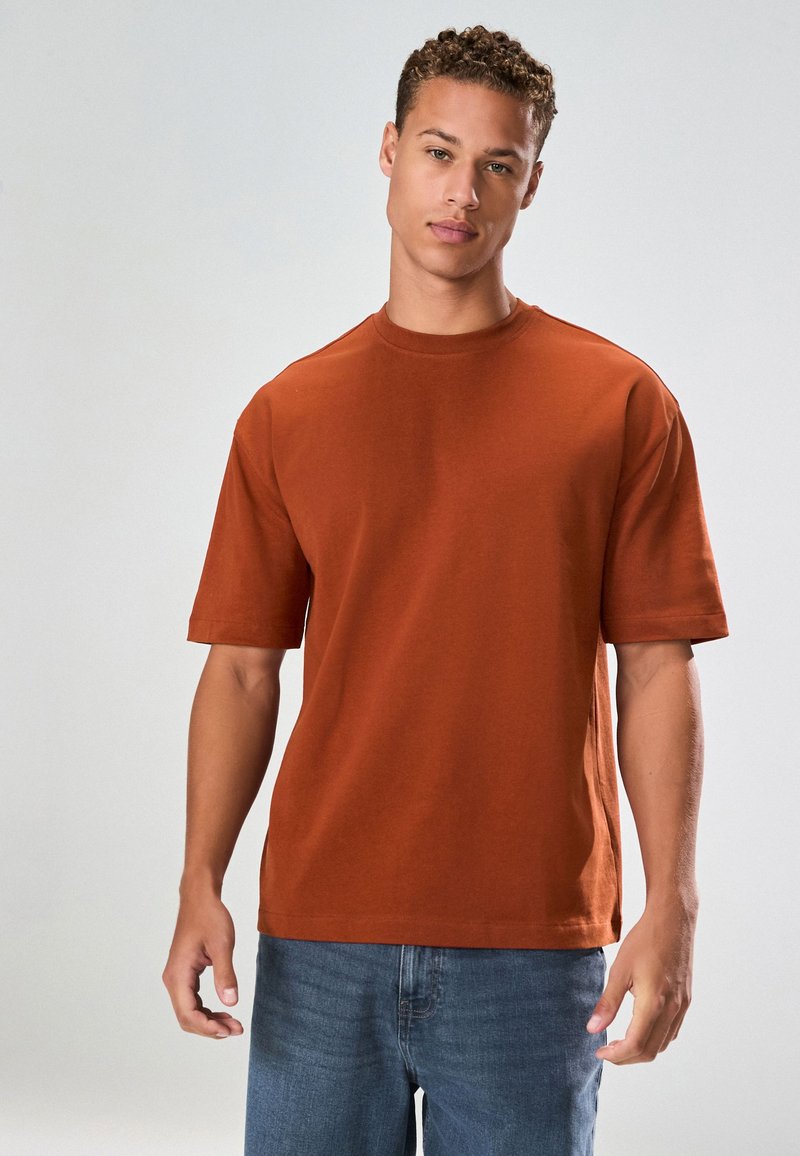 Kortärmad, rostfärgad T-shirt i en lätt bomullsblandning med rund halsringning och avslappnad passform. Bärs med blå denimjeans.