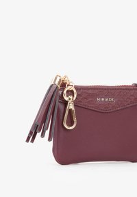 Borsetta in pelle bordeaux con finiture dorate, chiusura con zip e nappine decorative. Presenta un'area testurizzata e un logo in rilievo. Design compatto.