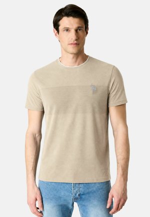 Uomo che indossa una maglietta beige a maniche corte con trama e jeans azzurri, in piedi davanti a uno sfondo chiaro e uniforme.