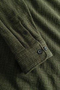 Groene, gestructureerde stof met een geknoopte manchet met twee zwarte knopen. Het patroon lijkt geweven met een rasterachtige textuur.