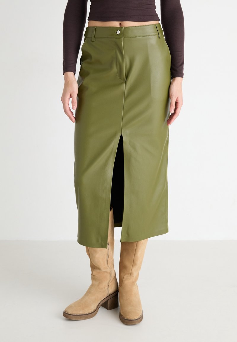 Jupe midi en cuir vert olive avec fente frontale portée avec des bottes hautes en daim couleur tan et un crop top à manches longues foncé.