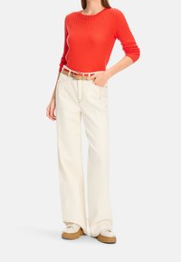 Haut à manches longues rouge à côtes associé à un pantalon large crème et une ceinture beige. Les chaussures sont crème avec une semelle épaisse beige. Tenue simple et décontractée.