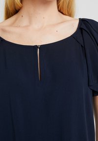 Marineblaues Blusenshirt mit Rundhalsausschnitt, das vorne über ein kleines Schlüssellochdetail verfügt. Kurze Puffärmel und eine weiche, glatte Stoffoberfläche.
