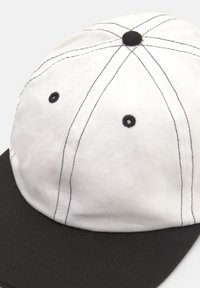 Fehér sapka fekete karimával, kontrasztos fekete varrással, lekerekített csúccsal és szellőző szemekkel. Nedvesebb textúra a felső anyagon.