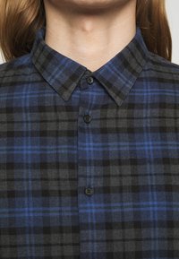 Rutig skjorta gjord av flanell. Färgerna inkluderar svart och olika nyanser av blått. Har en skjortkrage med knappar framtill och svarta knappar.