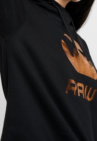 Persona vistiendo una sudadera con capucha negra con un logo de cobre brillante "RAW" y un gráfico estilizado en el pecho, mirando ligeramente de lado.