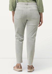 Pantaloni slim-fit grigio chiaro con orlo sfrangiato e due tasche posteriori, abbinati a sandali metallici marroni. Tessuto texturizzato visibile.