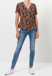 Blusa floral de manga corta, con escote en V profundo y forma peplum en burdeos, naranja y blanco. Combinada con jeans ajustados azules y zapatillas blancas.