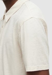 Polo beige clair en tissu doux et texturé. Il comporte un col, des manches courtes et des coutures visibles le long des bords des manches.