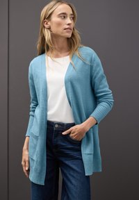 Ljusblå cardigan i mjukt material, med öppen fram, ribbade muddar och två framfickor, kombinerad med en vit skjorta och mörka jeans.