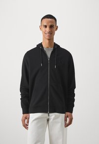Svart zip-up hettegenser med frontlommer, justerbar snor i hetten, og subtil logo detalj på brystet. Båret over en grå skjorte, kombinert med hvite bukser.