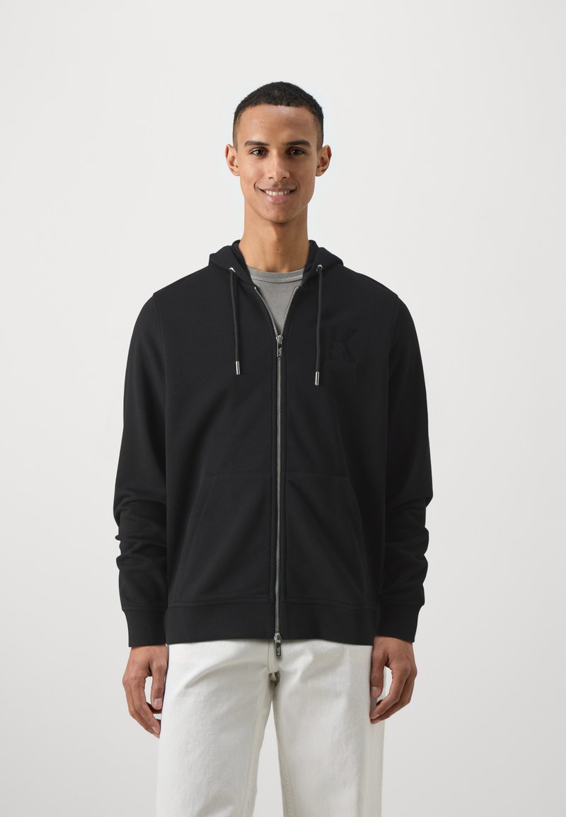 Svart zip-up hettegenser med frontlommer, justerbar snor i hetten, og subtil logo detalj på brystet. Båret over en grå skjorte, kombinert med hvite bukser.