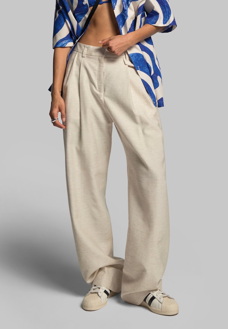 Personne portant un pantalon beige ample, des baskets blanches avec des bandes noires et une chemise ouverte à motifs bleu et blanc par-dessus un top bleu.