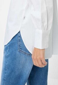 Witte langärmige shirt met knoopsluiting bij de manchetten, met een afgeronde zoom, gecombineerd met lichtblauwe spijkerbroek. Etiket zichtbaar aan de zijkant van het shirt.