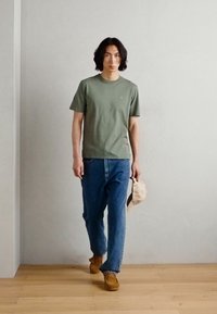 T-shirt en coton vert, jean en denim bleu ample, chaussures marron, tenant une casquette beige, se tenant devant un arrière-plan neutre avec un sol en bois.