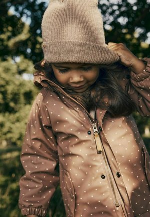 Giacca marrone a pois con zip e tasche laterali; caldo cappello di maglia beige; capelli che scendono fluenti. Ambientazione all'aperto con vegetazione sfocata.