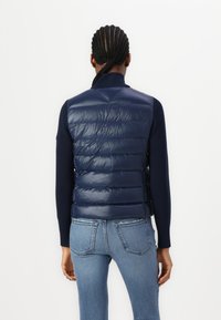Gilet imbottito blu navy con esterno lucido, colletto con zip e design trapuntato, abbinato a una maglietta a maniche lunghe blu navy e jeans blu.