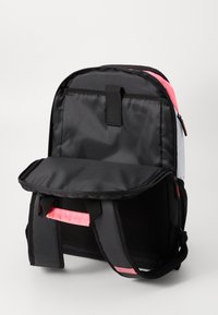 Mochila preta, rosa e branca com um compartimento traseiro aberto, alças de ombro acolchoadas e uma pega, apoiada numa superfície branca.