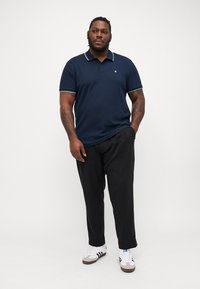 Polo de marină dintr-un material texturat, cu guler și accente pe mânecă în contrast alb, asociat cu pantaloni negri și adidași albi.