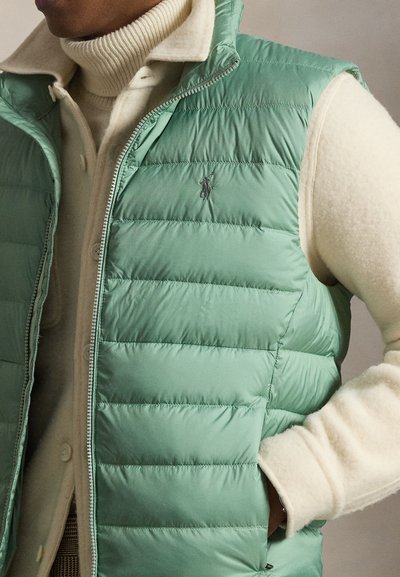 Άνδρας που φοράει ανοιχτοπράσινο μπουφάν χωρίς μανίκια με επένδυση πάνω από εκρού πουλόβερ με ζιβάγκο, με το χέρι στην τσέπη του μπουφάν, εμφανή φερμουάρ και λεπτομέρειες με κουμπιά.