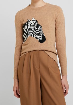 Beige sweater med sort og hvid zebrahoveddesign, båret sammen med brune, højtaljede bukser med vide ben, hænder afslappet langs siderne.