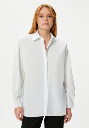 Camicia bianca a maniche lunghe con colletto, vestibilità comoda, texture liscia e orlo dritto. Indossata con pantaloni scuri.
