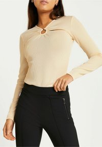 Beige Langarmoberteil mit einem Schlüssel-ausschnitt am Kragen, gerippte Textur; kombiniert mit hoch taillierten schwarzen Hosen mit Reißverschlusstaschen.